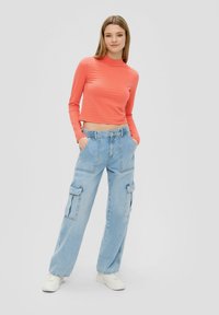 Koralju krāsas garām piedurknēm tops ar teksturētu rakstu, saskaņots ar gaiši ziliem bagāžnieka džinsiem, kuriem ir sānu kabatas. Modelis valkā baltas sporta kedas.