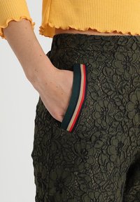 Zielone koronkowe spodnie z kwiecistym wzorem, z kieszenią wykończoną w paski w kolorze czerwonym, kremowym i granatowym. Teksturowany materiał, dopasowany krój.