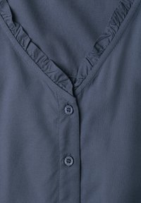 Col de chemise gris foncé avec petite bordure à volants et deux boutons ronds visibles avec texte sur le tissu.