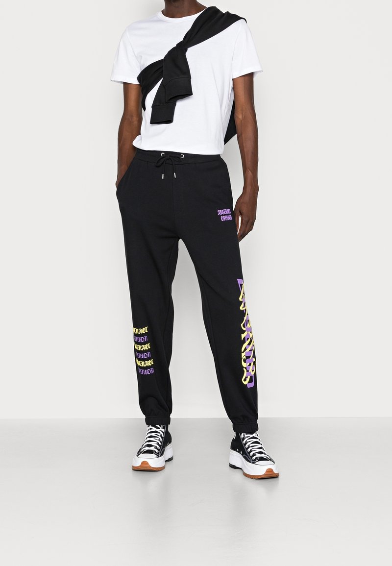 Svarta sweatpants med lila och gula textgrafiker, dragsko i midjan och elastiska muddar, kombinerat med en vit T-shirt och svarta skor.
