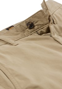 Närbild av beige byxmidja med brun knapp, dragkedja delvis öppen och synlig svart och orange BOSS-logotyp.