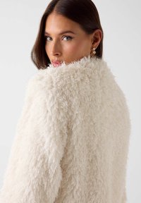 Witte nepbontjas met een gestructureerd, pluizig oppervlak. Voorzien van een hoge kraag en een open voorkant, gedragen over de schouders.