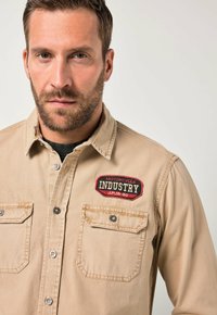 Beżowa koszula zapinana na guziki, bawełniana tkanina, dwie kieszenie na piersi, czerwona i czarna naszywka z napisem "MOTORCYCLE INDUSTRY JP 18-80".