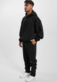 Schwarzes Hoodie- und Sweatpants-Set aus weichem Material. Verfügt über eine Fronttasche und Bündchen an den Ärmeln. Kombiniert mit weißen Turnschuhen.