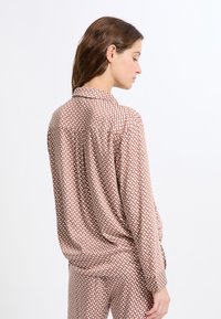 Bluzka z długim rękawem wykonana z jasnoróżowego materiału z geometrycznym wzorem w ciemnoróżowym i białym kolorze. Posiada kołnierzyk, luźny krój oraz zdobienie na guziki.