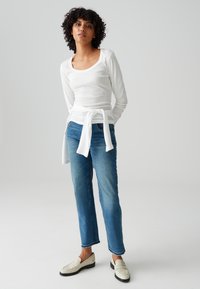 Frau mit lockigem Haar, die ein weißes, langärmliges Oberteil trägt, das an der Taille gebunden ist, blaue Jeans und weiße Loafers, steht vor einem einfarbigen Hintergrund.