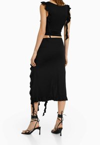 Schwarzes zweiteiliges Outfit mit einem engen Oberteil mit Rüschensäumen und einem Midirock mit Rüschendetails. Der Rock hat einen seitlichen Schlitz und Bindebänder.