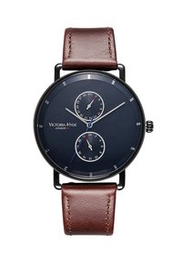 Cadran bleu marine rond avec deux sous-cadrans, aiguilles argentées, boîtier noir et bracelet en cuir marron. Logo "Victoria Hyde London" affiché.