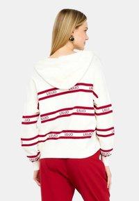 Felpa bianca con strisce orizzontali rosse e logo "LIUJO". Presenta un cappuccio regolabile e una vestibilità comoda. Abbinata a pantaloni rossi.