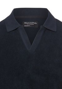 Polo bleu foncé avec col et ouverture en V, montrant une étiquette Marc O'Polo et une broderie discrète du logo sur la poitrine.