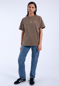 Camiseta de algodón marrón con un corte relajado, mangas cortas y un pequeño detalle de logo. Combinada con jeans de mezclilla azul claro y zapatillas negras.