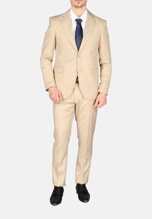 Man met beige tweeknopssuit, wit overhemd, marineblauwe stropdas en zwarte nette schoenen, staande voor een effen achtergrond.