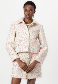 Veste en tweed pastel avec un design à col et boutons sur le devant, accompagnée d'une jupe mini assortie présentant des coutures visibles et une texture.