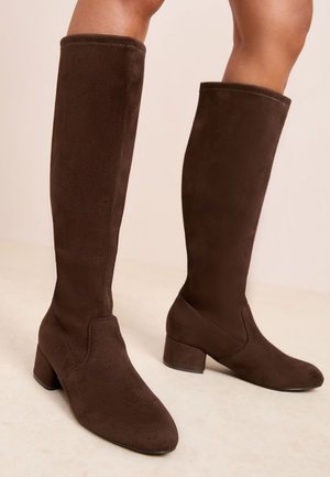 Bruine suède kniehoge laarzen met blokhakken, gedragen op blote benen, getoond tegen een neutrale achtergrond.