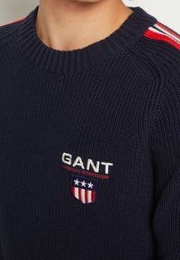 Marineblauer Strickpullover mit geripptem Halsausschnitt, der rote und weiße Streifenakzente auf den Schultern und ein besticktes Logo auf der Brust aufweist.