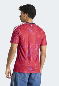 Le maillot de sport rouge à manches courtes présente un motif abstrait violet, un col rond et trois bandes blanches sur les épaules.