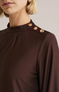 Rjava bluza z visokim vratom, s pliseji in tremi kovinskimi gumbi na ramenu, narejena iz gladkega materiala.