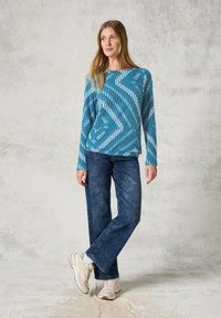Turkusowy sweter z białymi skośnymi paskami, o luźnym kroju. Połączony z ciemnoniebieskimi spodniami typu wide-leg i neutralnymi trampkami.