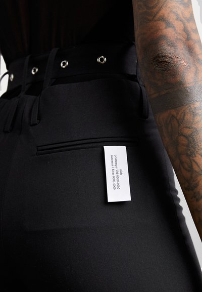 Black trousers made of smooth fabric, featuring a high waist, large eyelet detailing, and a small tag on the back pocket.