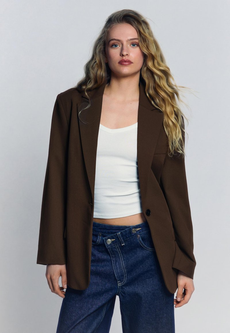 Hosszú hullámos szőke hajú nő barna, túlméretezett zakóban, fehér rövid felsőben és magas derekú kék farmerral, szürke háttér előtt állva.