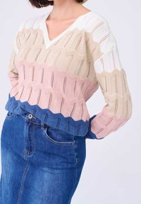 Femme portant un pull tricoté multicolore avec des vagues blanches, beiges, roses et bleues, associé à une jupe en denim bleu taille haute.