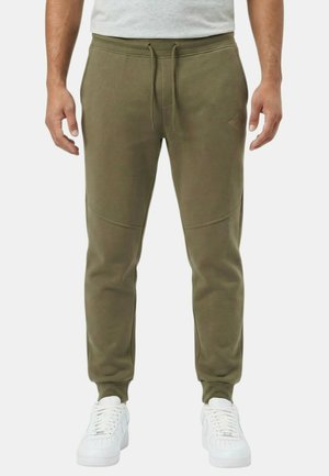 Homme portant un pantalon de jogging vert olive à taille élastique avec cordon et des baskets blanches, debout devant un fond uni.