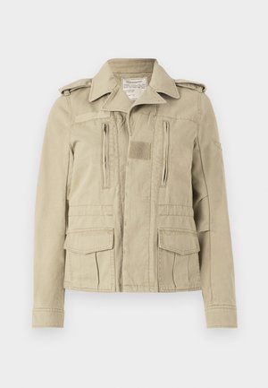 Jachetă din bumbac de culoare bej, stil militar, cu închidere cu butoane, două buzunare laterale cu fermoar și buzunare cu clapetă pe lateral. Are epauleți și un guler texturat.