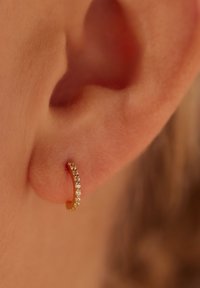 Gouden oorbel met een rij kleine heldere kristallen langs de voorkant, gedragen aan het oor van een persoon in close-up. Gladde textuur, ronde vorm.