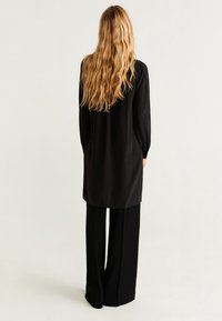 Tunique noire à manches longues avec une coupe droite, associée à un pantalon noir à jambes larges. Tissu lisse avec un léger éclat, sans motifs visibles.