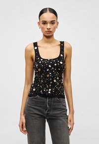 Frau mit zurückgekämmtem Haar, trägt ein schwarzes ärmelloses Oberteil, verziert mit mehrfarbigen Pailletten, und dunkelgraue High-Waist-Jeans.