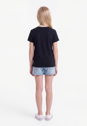 Kind mit langen blonden Haaren, trägt ein schwarzes T-Shirt, helle Jeansshorts und weiße Sneakers, steht mit dem Rücken zur Kamera vor einem weißen Hintergrund.