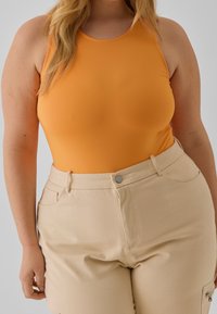 Bodysuit sin mangas de color naranja con una textura suave, combinado con pantalones de tiro alto de color beige claro que cuentan con bolsillos delanteros y herrajes metálicos.