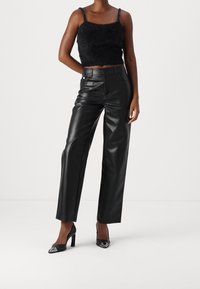 Top corto in pelliccia sintetica nera con spalline sottili, abbinato a pantaloni in pelle nera a vita alta. Scarpe con tacco alto nere con dettagli argentati.