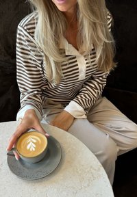 Ein Latte mit Blattdesign in einer hellblauen Tasse auf einem grauen Teller, gehalten von einer Person in einem braun-weiß gestreiften Shirt, die an einem Marmortisch sitzt.
