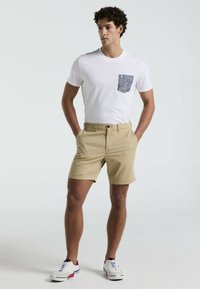 Weißes Kurzarmhemd mit gemustertem Taschen; beige Shorts; das Modell steht mit Händen in den Taschen und trägt weiße Sneaker mit roten Akzenten.