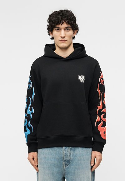 Tânăr purtând un hanorac negru cu flăcări albastre pe mâneca stângă, flăcări roșii pe mâneca dreaptă și un mic logo alb pe piept.