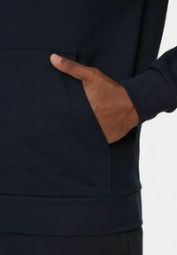 Felpa blu navy con tasca frontale e polsini a coste. Il tessuto appare morbido con una consistenza liscia. Mano posizionata sulla tasca.