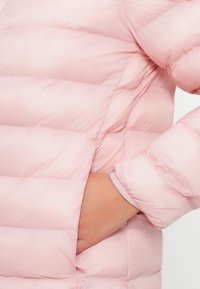 Svetlo roza puffer jakna z prešitimi deli, z zadrgo na žepu in rebrastimi manšetami. Material se zdi gladek z mehkobo.