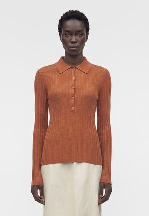 Γυναίκα φοράει μακρυμάνικη, ριμπ πλεκτή, σκουροκόκκινη ζακέτα πόλο με κουμπιά και ανοιχτό μπεζ φούστα σε ουδέτερο φόντο.