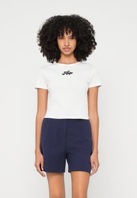 Balta cropped t-kreklus ar īsām piedurknēm, uz kura redzams melns "Hugo" teksts. Saskaņots ar tumši ziliem piegulošiem šortiem no gludas auduma ar strukturētu piegriezumu.