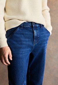 Jeans de mezclilla azul oscuro con un diseño de bolsillo delantero y un ligero desvanecimiento en el muslo, combinados con un suéter de punto crema.