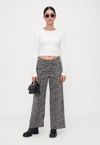 Femme avec des lunettes de soleil noires, un haut court à manches longues blanc, un pantalon large à imprimé zèbre, des bottes noires, tenant un petit sac à main noir.