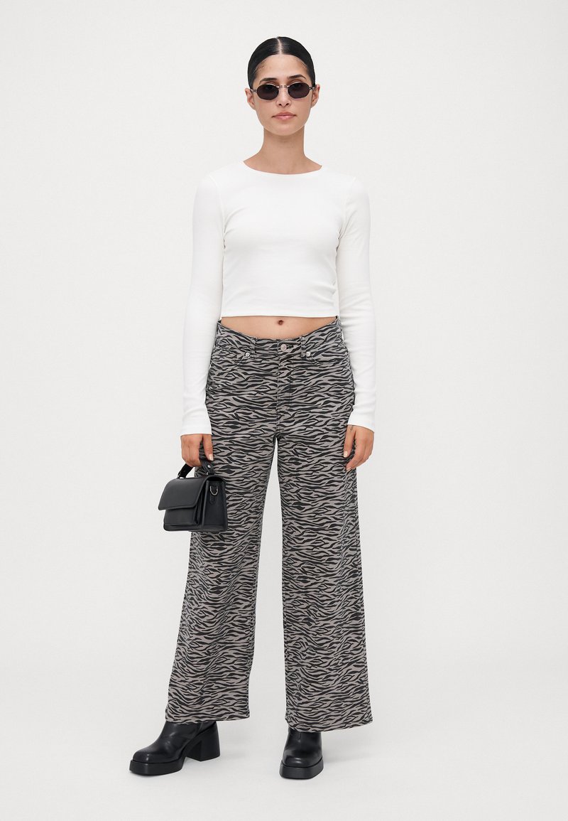 Femme avec des lunettes de soleil noires, un haut court à manches longues blanc, un pantalon large à imprimé zèbre, des bottes noires, tenant un petit sac à main noir.