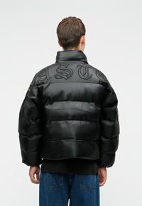 Pessoa a usar um casaco acolchoado preto com letras elevadas na parte superior das costas, combinado com jeans azuis, vista de trás.