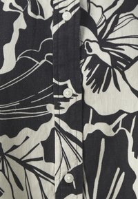 Chemise à motif floral noir et blanc. Présente une patte de boutonnage avec des boutons blancs, confectionnée dans un tissu texturé. Le design est audacieux et contrasté.