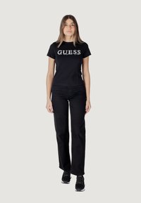 Zwarte katoenen T-shirt met "GUESS" logo, gecombineerd met zwarte denim jeans. Model draagt zwarte sneakers, met een rechte pijp en minimale details.