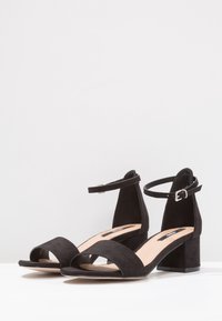 Schwarze Wildleder-Sandalen mit Blockabsatz, Knöchellasche und offenen Zehen, die auf einer weißen Fläche vor einem schlichten weißen Hintergrund präsentiert werden.