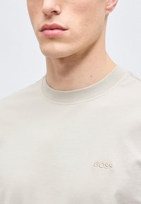 Κοντινό πλάνο ενός ατόμου που φοράει ανοιχτό μπεζ μπλουζάκι με στρογγυλή λαιμόκοψη και κεντημένο λογότυπο "BOSS" στο αριστερό μέρος του στήθους.