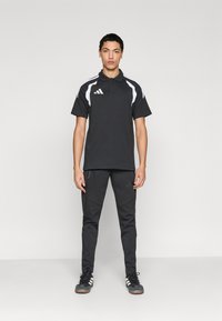 Μαύρο αθλητικό πόλο με λευκές λεπτομέρειες και λογότυπο με τρεις ρίγες, συνοδευόμενο από μαύρο παντελόνι με στενή εφαρμογή και υφασμάτινα πάνελ με λογότυπο.