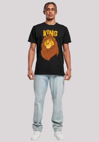 Črna bombažna majica s grafikami stilizirane glave leva in napisom "KING" v debelih rumenih črkah. Model nosi svetle jeans hlače in bele superge.